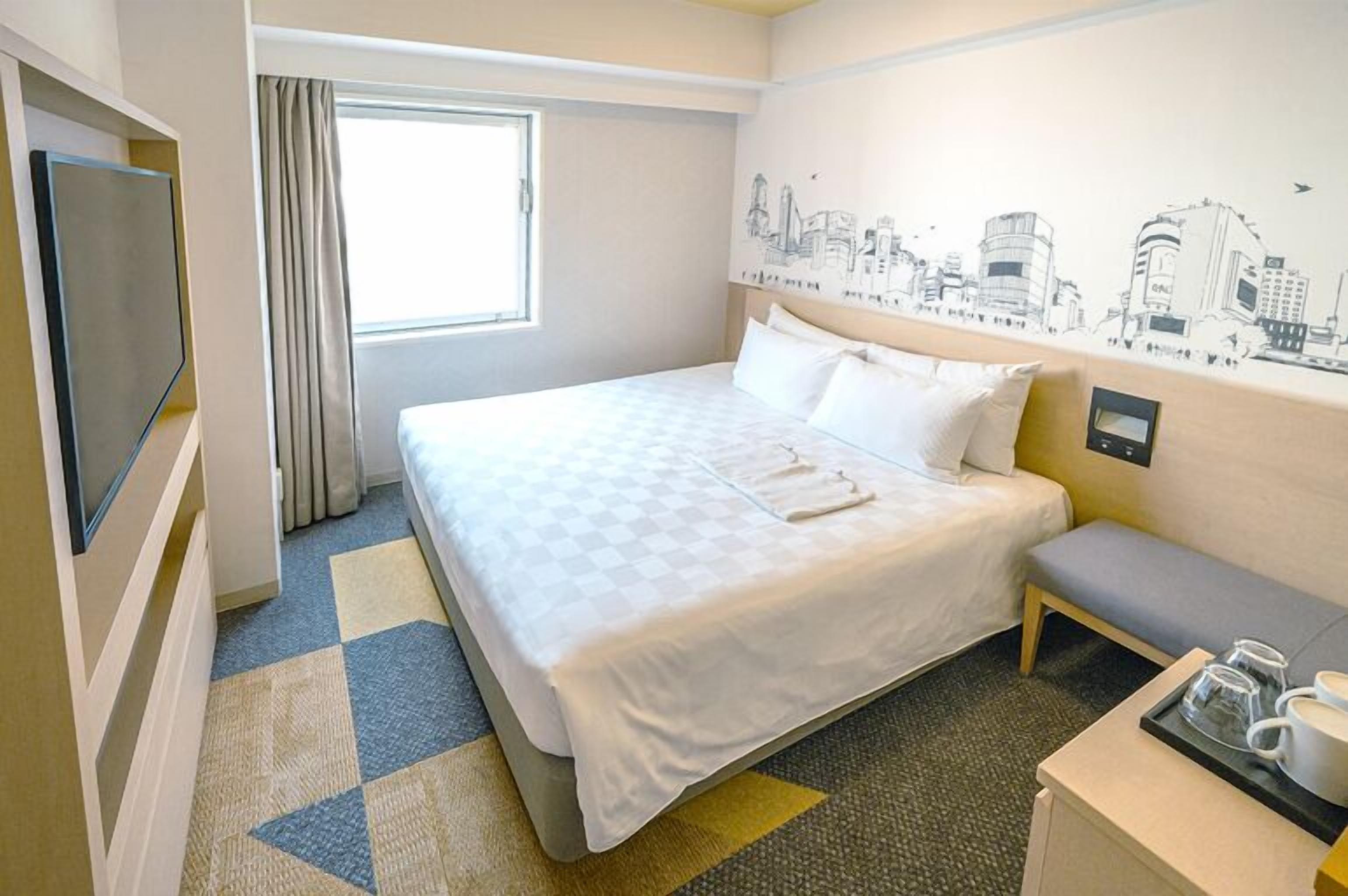 Habitación de hotel con cama, un banco, televisión, ventana con cortinas y un mural de paisaje urbano.