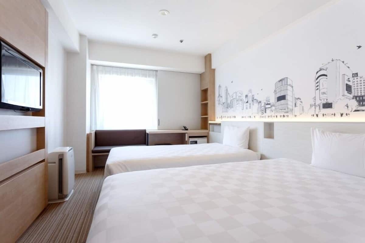 Ein Hotelzimmer mit zwei Betten, einem Schreibtisch, einem Sessel, einem Fernseher und einem Wandbild mit Stadtansicht.