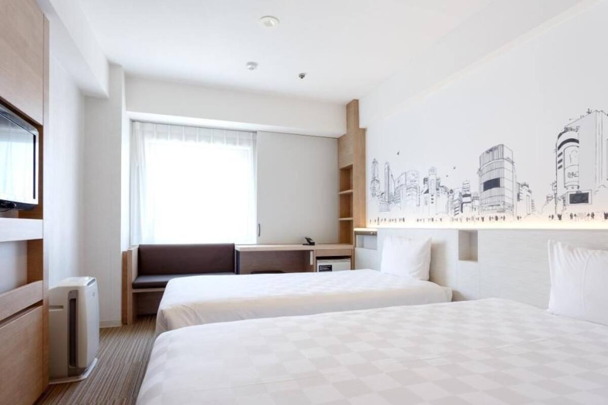 Ein Hotelzimmer mit zwei Betten, einem Schreibtisch, einem Stuhl, einem Heizkörper und einem Wandbild mit Stadtansicht.