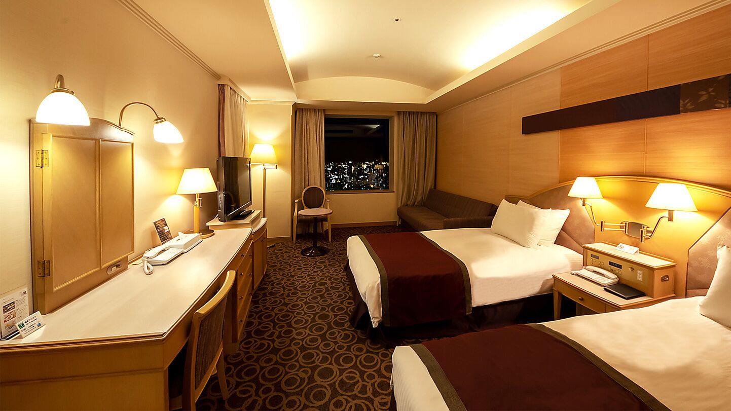 A hotel room with two beds, a desk, a chair, a TV, and a window with a city view.