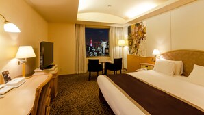 A hotel room with a bed, desk, chair, TV, and a view of a cityscape at night. - Ginza Creston (Tokyo)