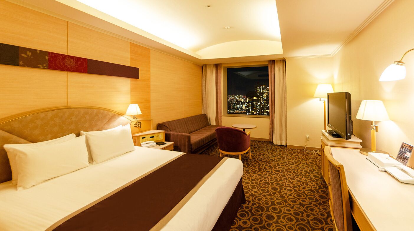 A hotel room with a large bed, a desk with a TV, a chair, a window with a city view, and a lamp.