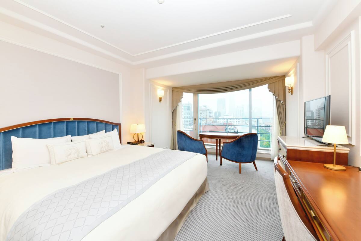 Quarto de hotel com uma cama grande, uma escrivaninha com televisão, duas cadeiras e vista da cidade através de amplas janelas.
