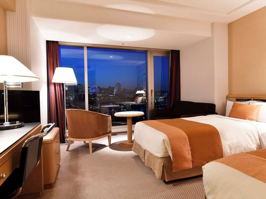 A hotel room with a large bed, a desk with a TV, a chair, a small table, and a view of the city at night.