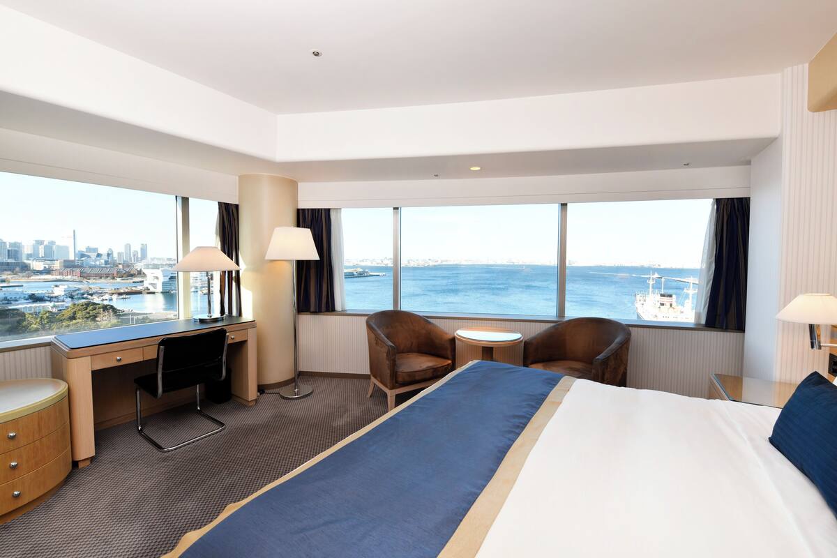 Quarto de hotel com uma cama grande, uma mesa, duas cadeiras, uma mesinha e vista para a cidade e para o mar.