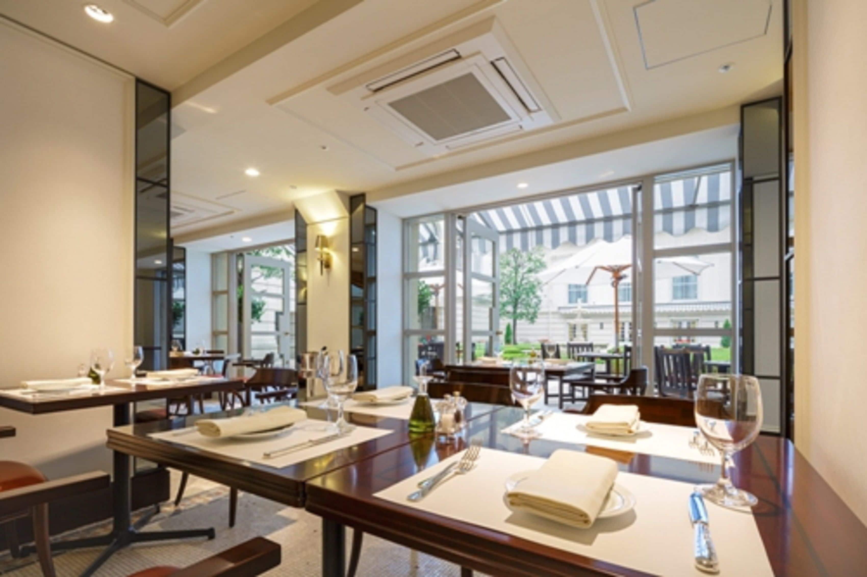 A modern dining area with tables set for two, large windows offering a view of the outdoors, and a clean, contemporary interior design.