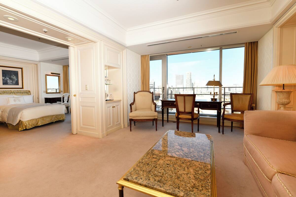 Quarto de hotel com cama, área de estar com mesa e cadeiras, varanda com vista para a cidade e uma janela grande.