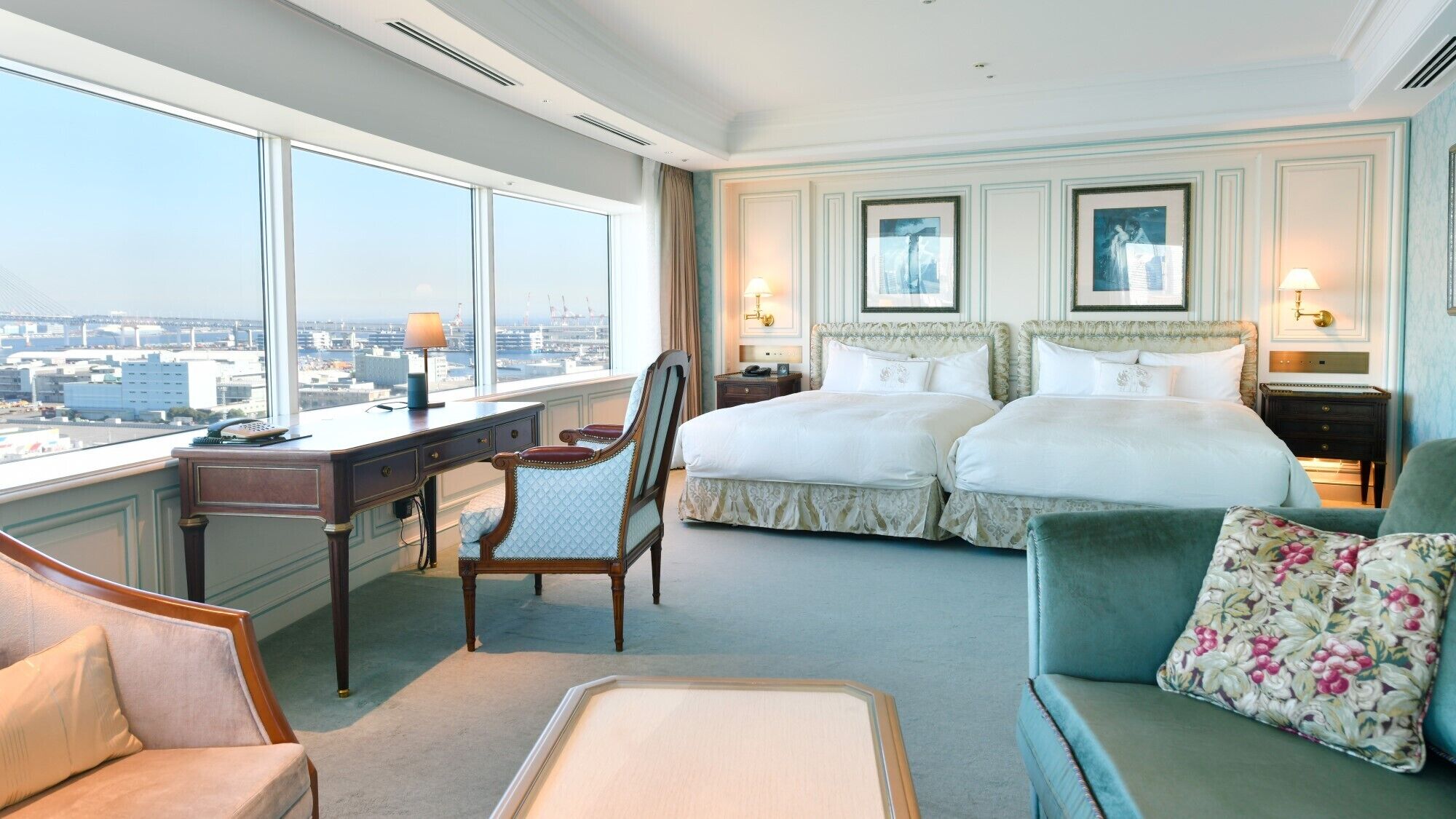 A hotel room with a city view, a desk with a lamp, two beds, a chair, and a sofa.