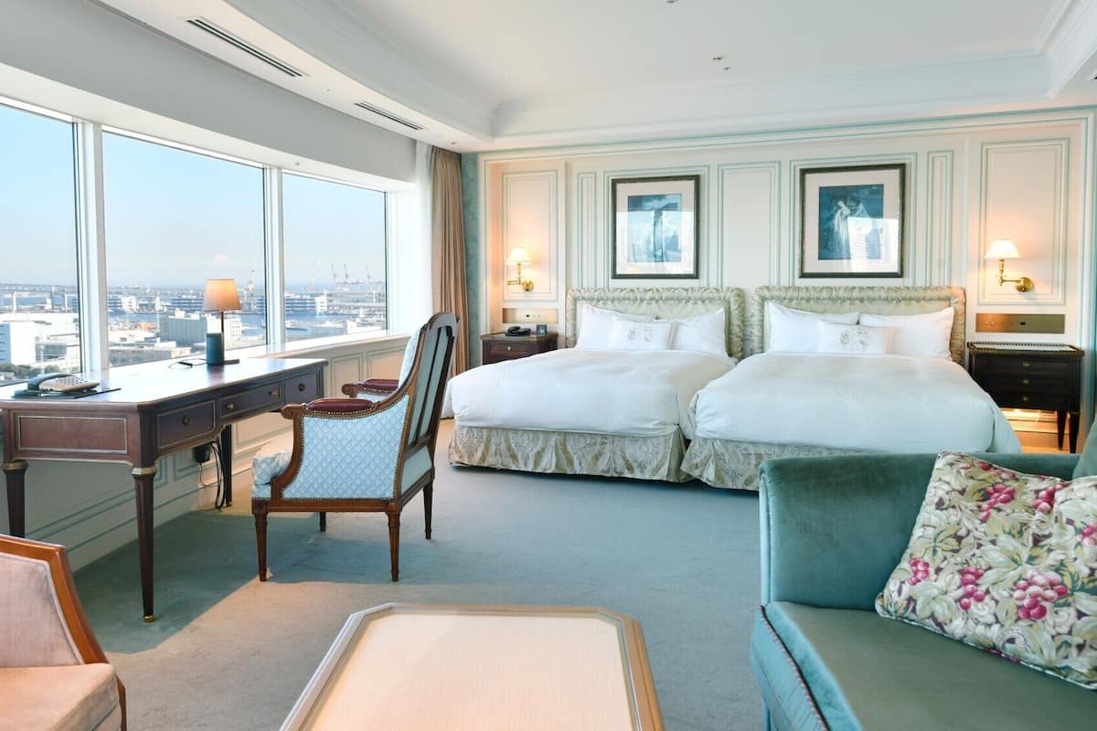 Quarto de hotel com vista para a cidade, uma escrivaninha com luminária, duas camas, uma cadeira e um sofá.