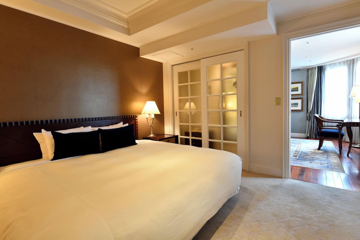 Quarto de hotel com uma cama grande, mesa de cabeceira com luminárias, porta que dá para uma sala de jantar e vista para a sala de jantar através de uma porta de vidro.