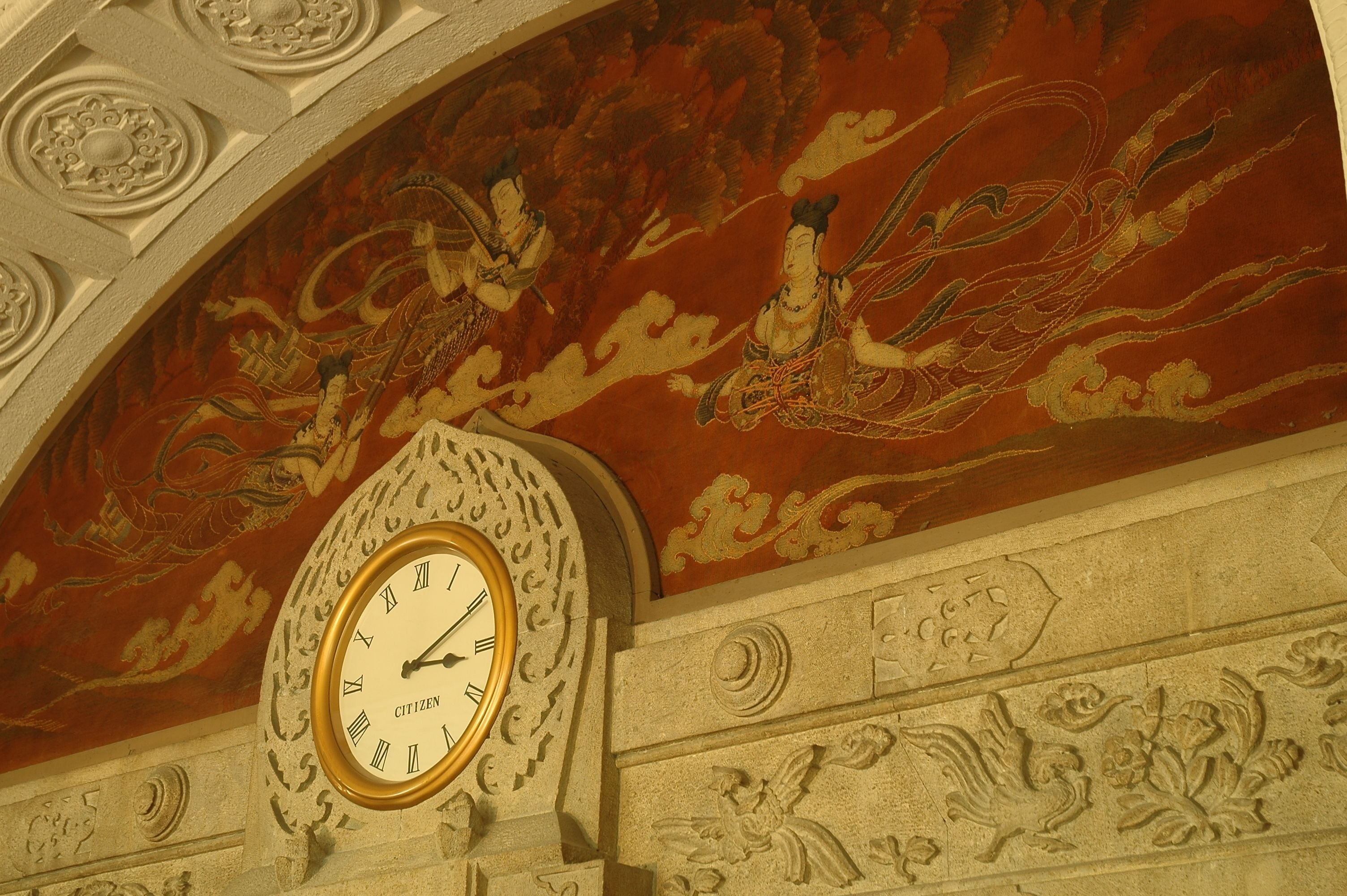 A clock with ornate carvings and a mural of mythological figures on the wall.