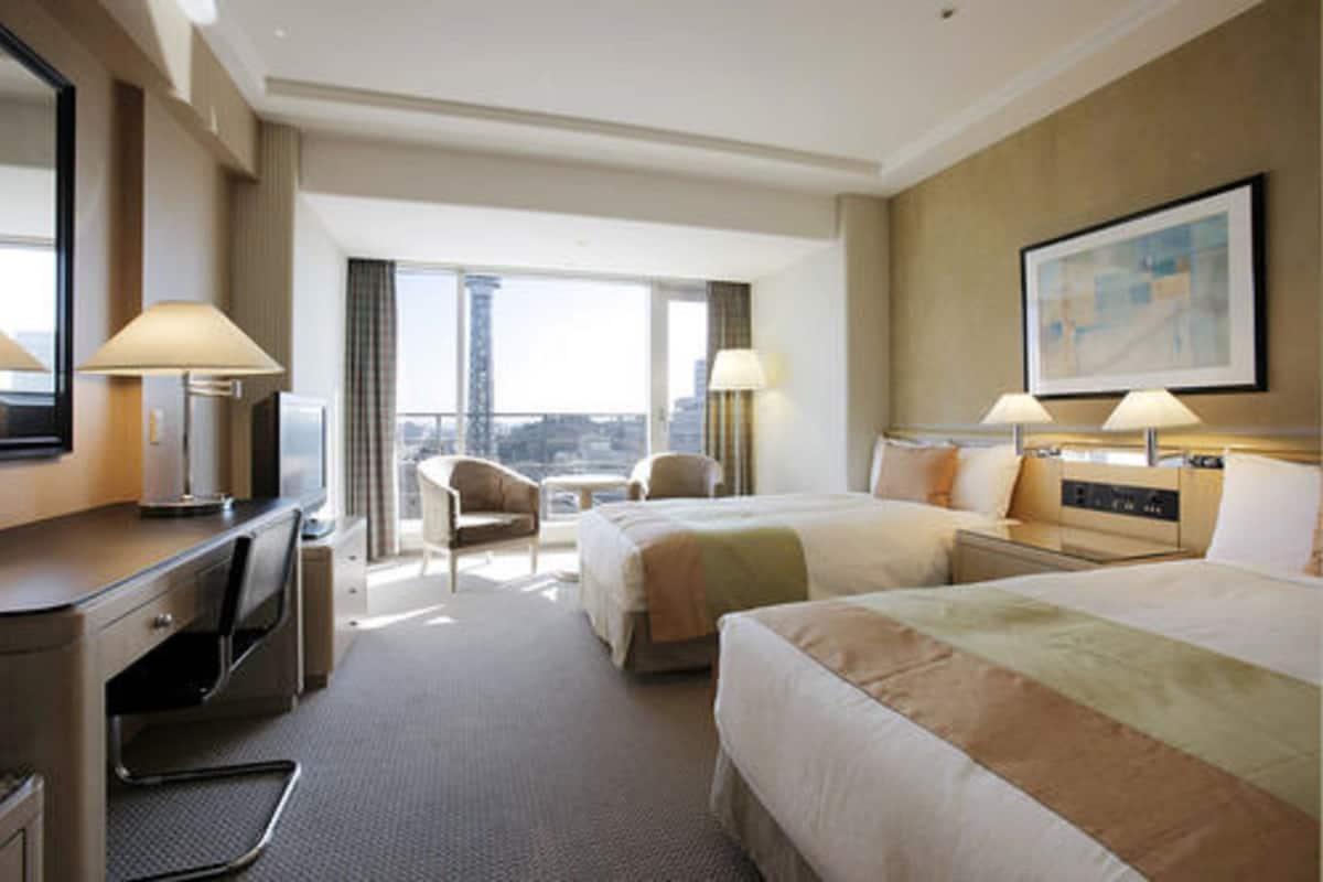 Quarto de hotel com duas camas, uma mesa, uma cadeira e uma janela grande com vista para a cidade.