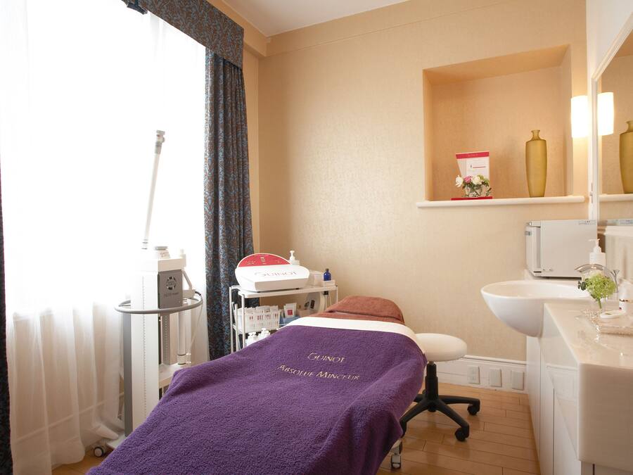 A room with a bed covered in a purple blanket, a white sink, and a shelf with decorative items.