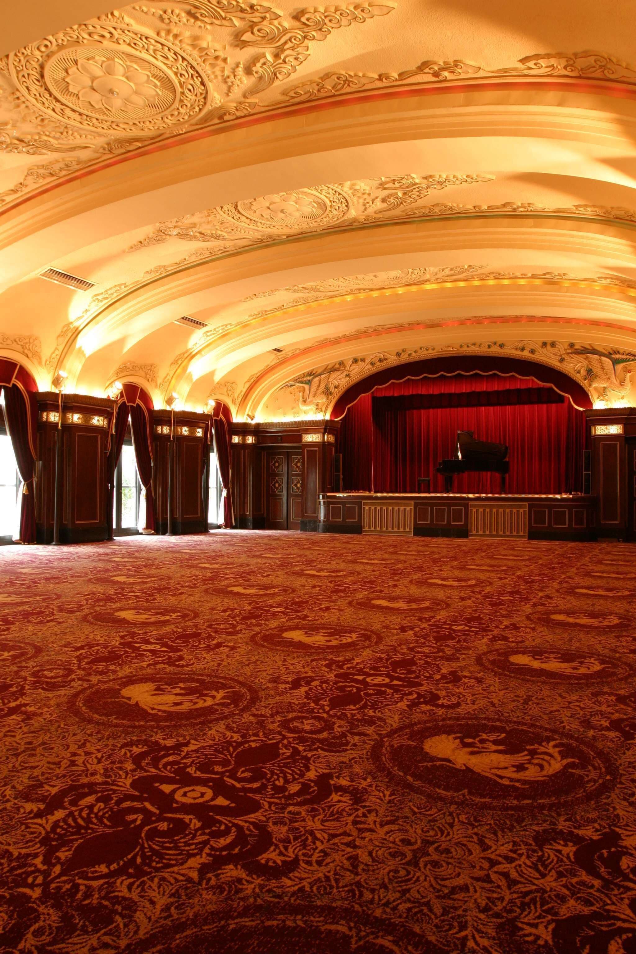Un vaste hall doté d’un plafond richement décoré et de rideaux rouges, avec une scène où se trouve un magnifique piano à queue.
