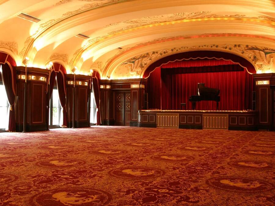 Un vaste hall doté d’un plafond richement décoré et de rideaux rouges, avec une scène où se trouve un magnifique piano à queue.