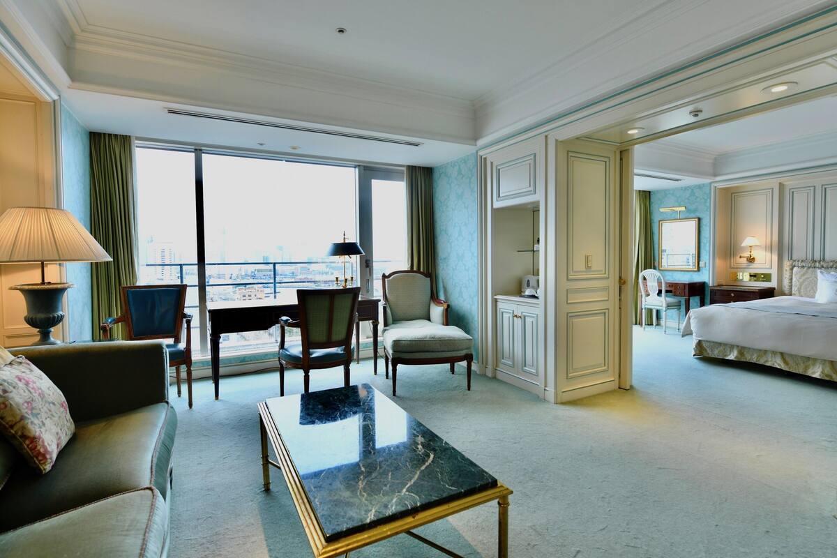 Quarto de hotel com uma cama grande, área de estar com sofá e cadeiras, mesa de jantar e vista da cidade por grandes janelas.