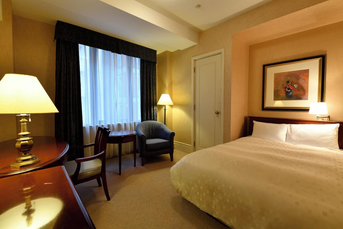 Quarto de hotel com cama, duas cadeiras, uma escrivaninha, abajur e um quadro na parede.