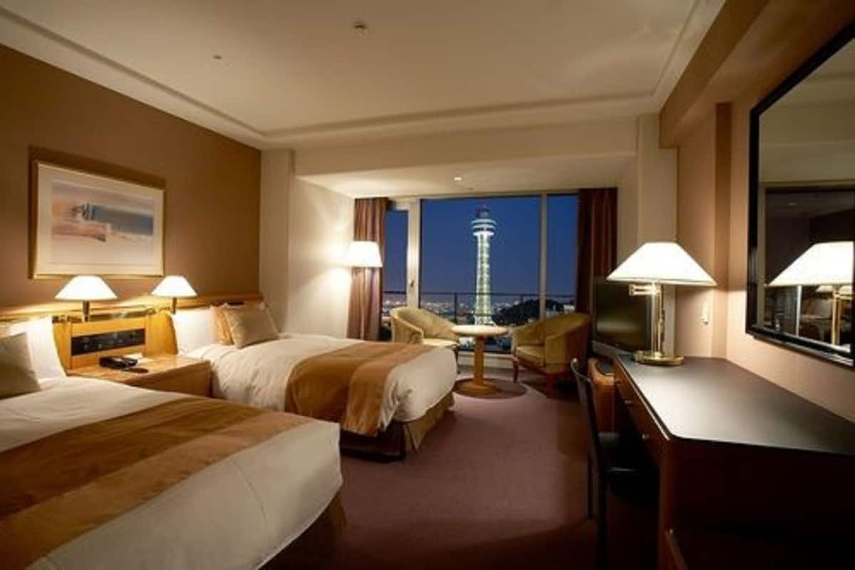 Quarto de hotel com duas camas, uma escrivaninha, uma cadeira e vista para uma torre à noite.