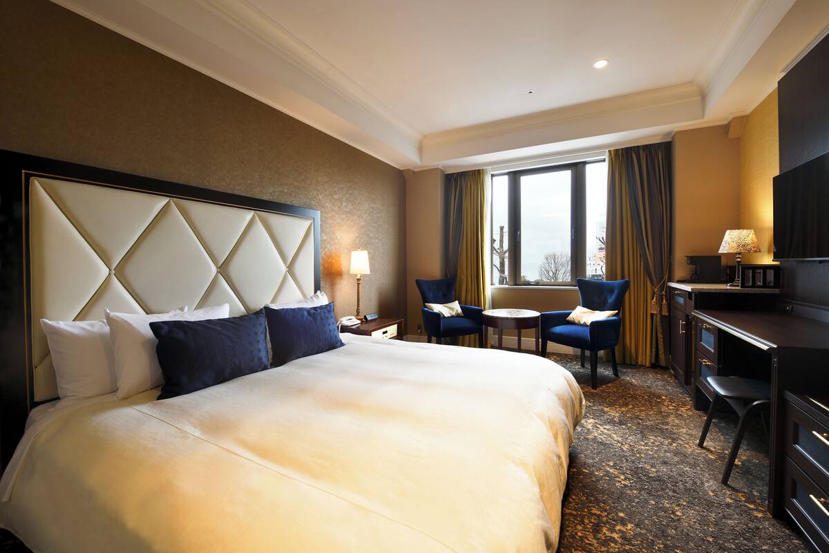 Quarto de hotel com uma cama grande, uma escrivaninha, uma cadeira e vista para a cidade pela janela.