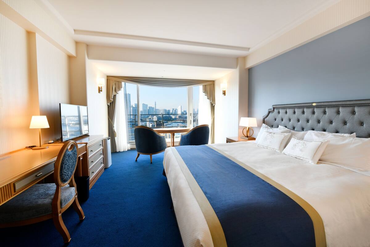 Quarto de hotel com uma cama grande, uma escrivaninha, uma cadeira e vista para o horizonte da cidade.