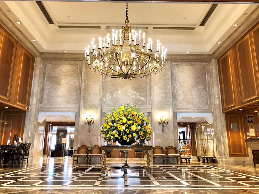 A spacious lobby with a grand chandelier, a floral centerpiece, and a checkered marble floor.