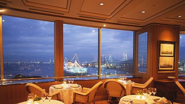 Un restaurant avec des tables rondes et des chaises en osier, de grandes fenêtres offrant une vue sur un pont et le paysage urbain au crépuscule.