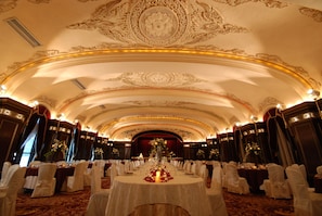 Um amplo salão de banquetes com ornamentos elaborados no teto, mesas redondas preparadas para refeições e cadeiras brancas.