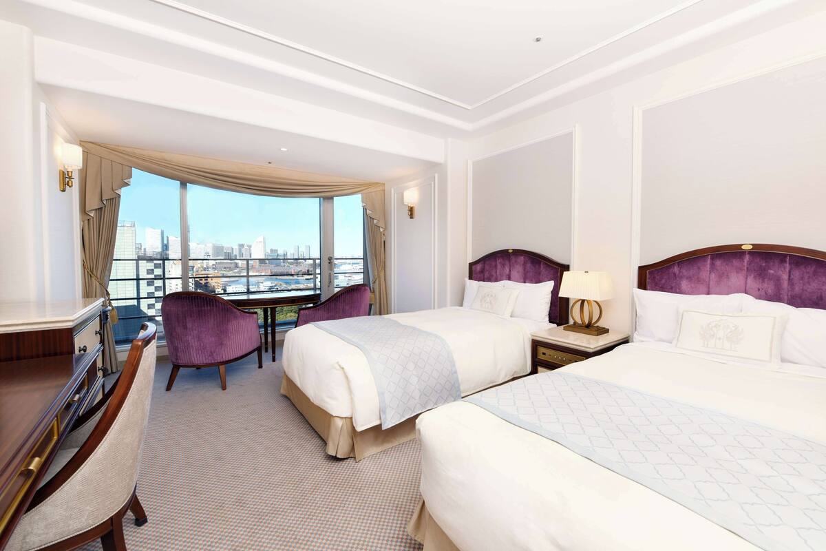 Quarto de hotel com duas camas, uma mesa, uma cadeira e vista para a paisagem urbana.