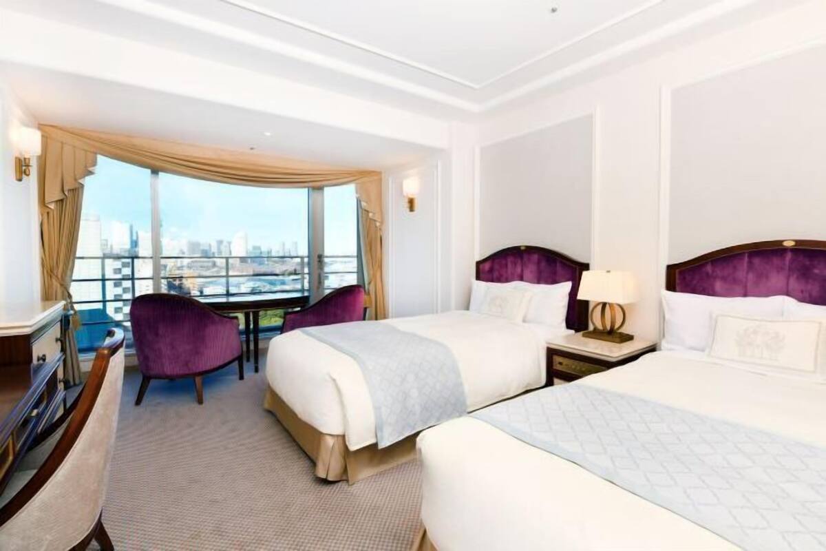 Quarto de hotel com duas camas, uma escrivaninha, uma cadeira e uma janela grande com vista para a cidade.
