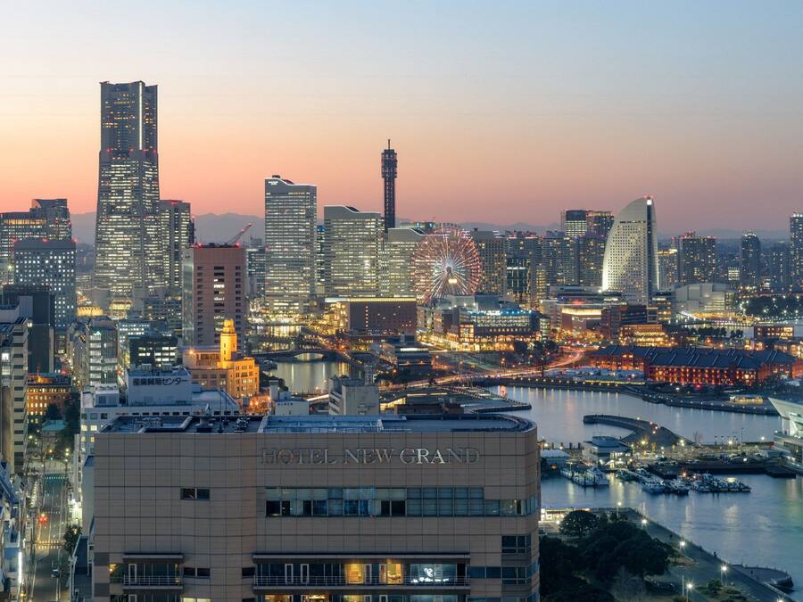 Stadsgezicht met een opvallend hotel genaamd "HOTEL NEW GRAND" op de voorgrond, omringd door wolkenkrabbers en een havengebied.