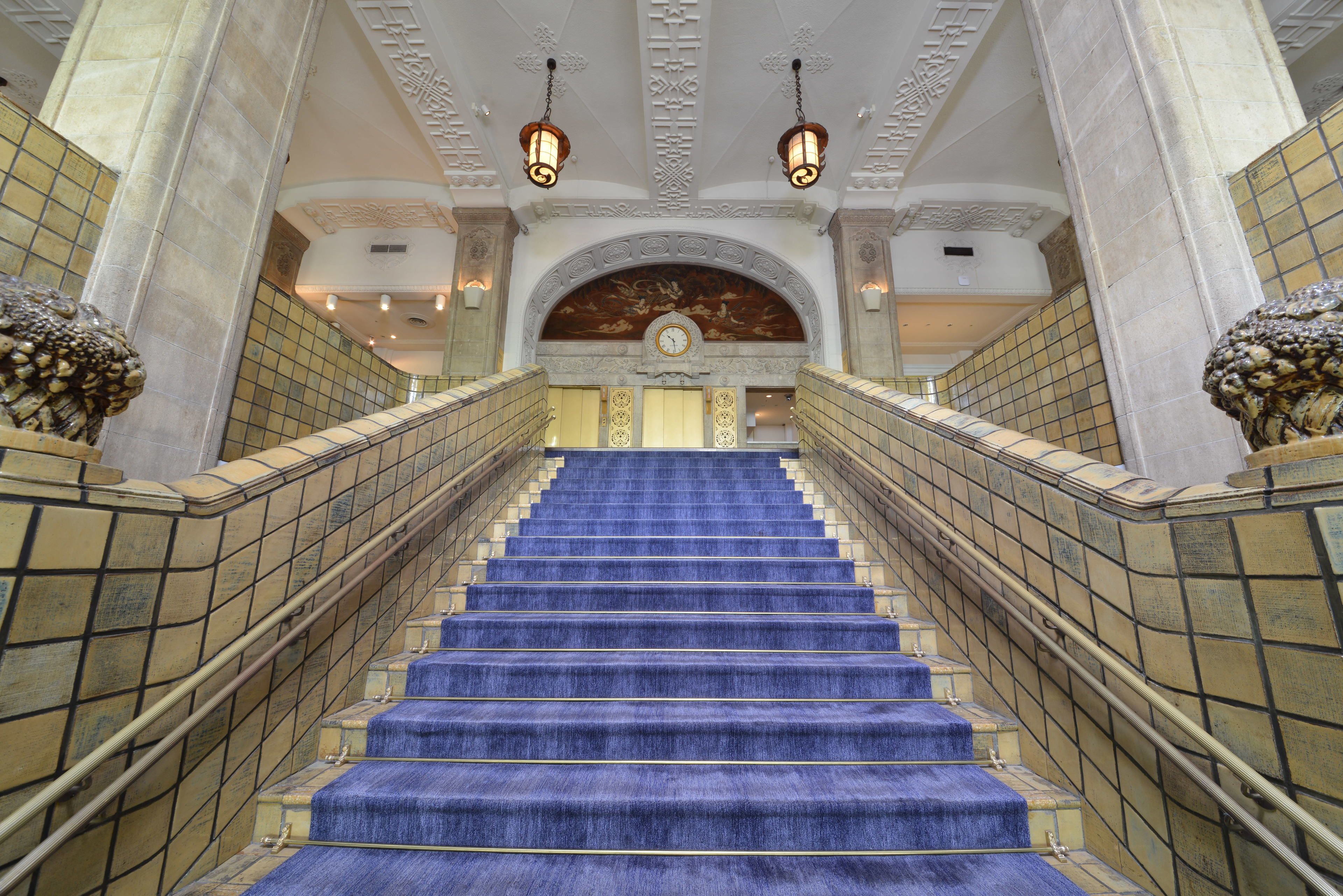 A grand staircase with blue carpeted steps, flanked by ornate columns and a decorative ceiling with hanging lamps.
