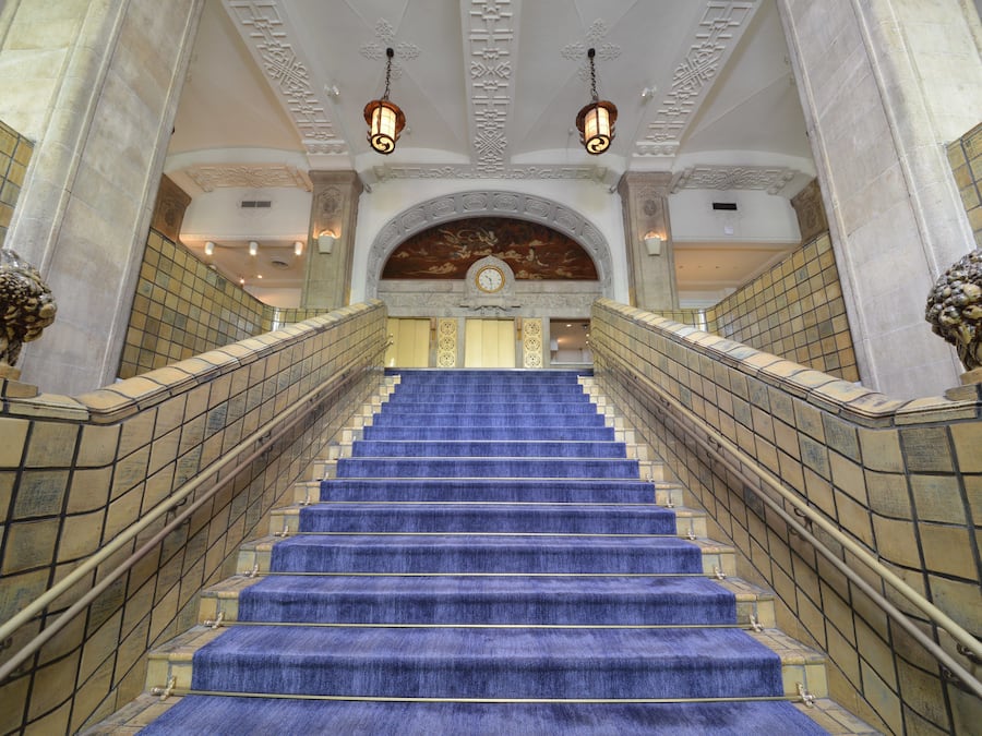 A grand staircase with blue carpeted steps, flanked by ornate columns and a decorative ceiling with hanging lamps.