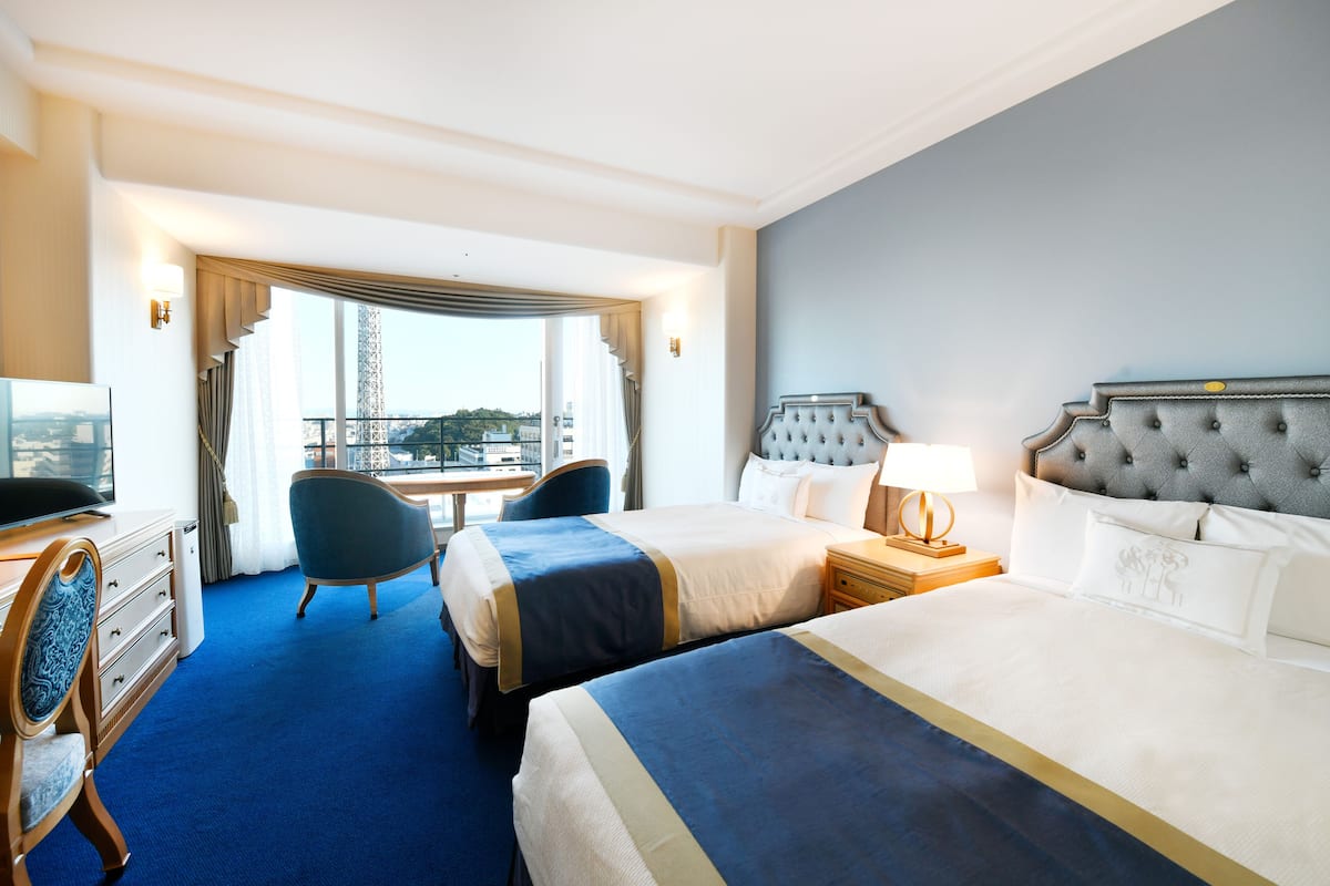 Quarto de hotel com duas camas, uma escrivaninha, uma cadeira e vista para a paisagem urbana.