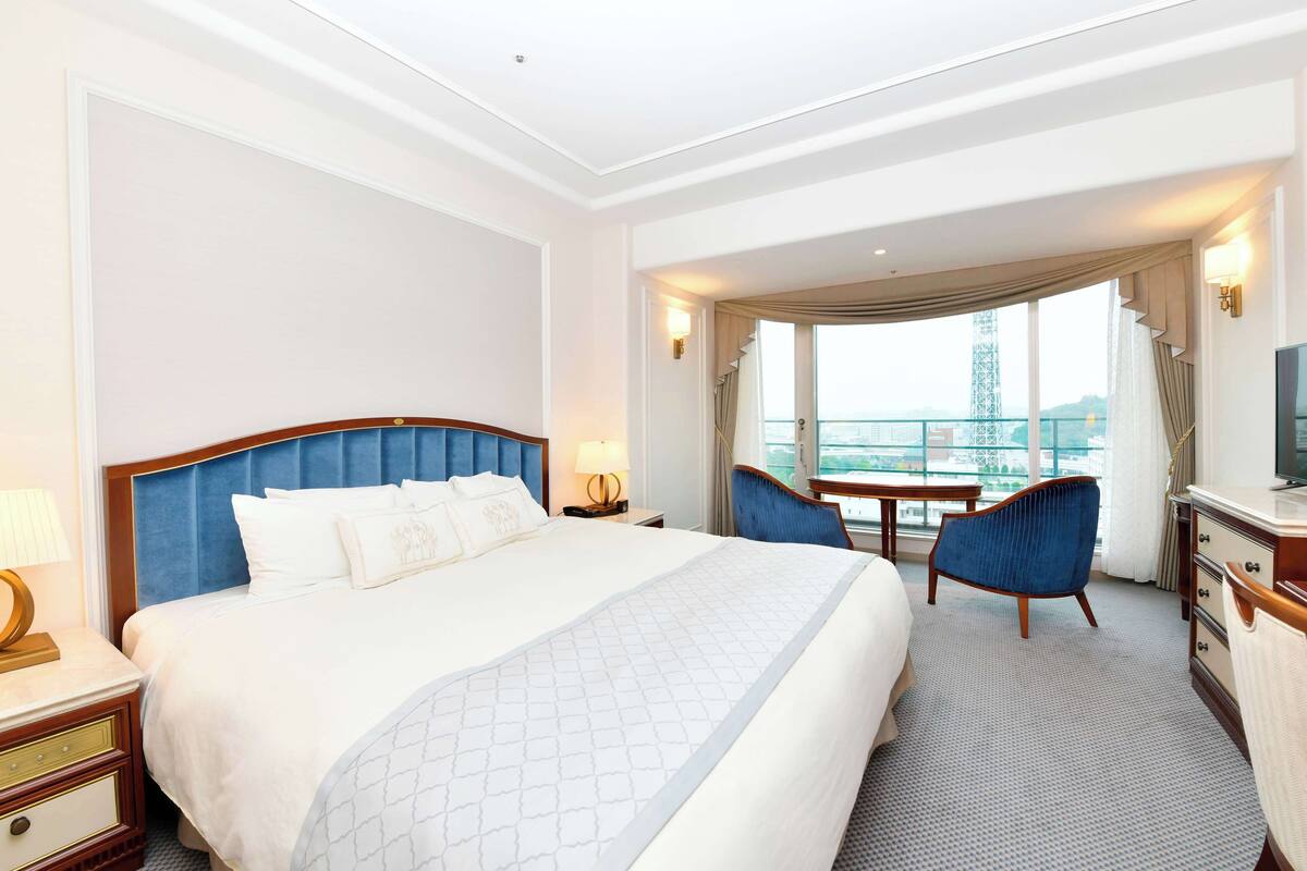 Quarto de hotel com uma cama grande, uma escrivaninha, uma cadeira e vista para uma torre.