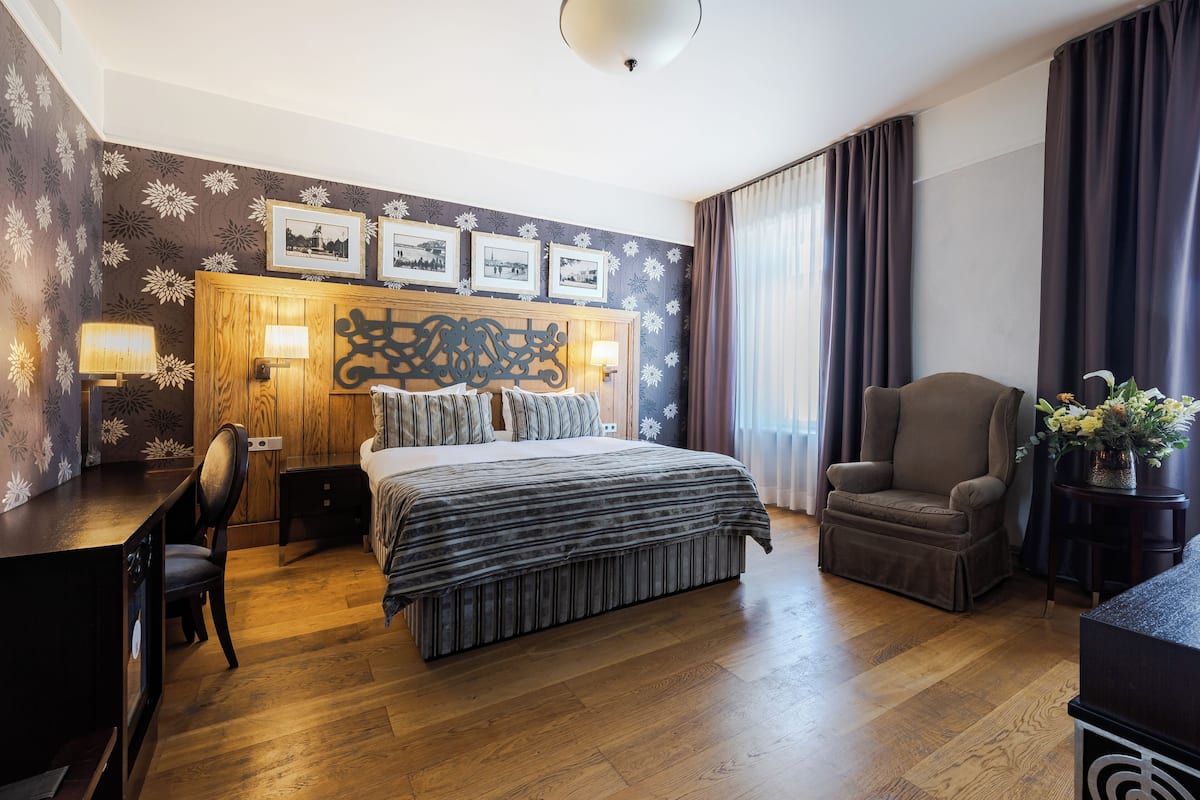 A hotel room with a wooden floor, a bed with a striped bedspread, a desk with a chair, a sofa, and a large window with curtains.