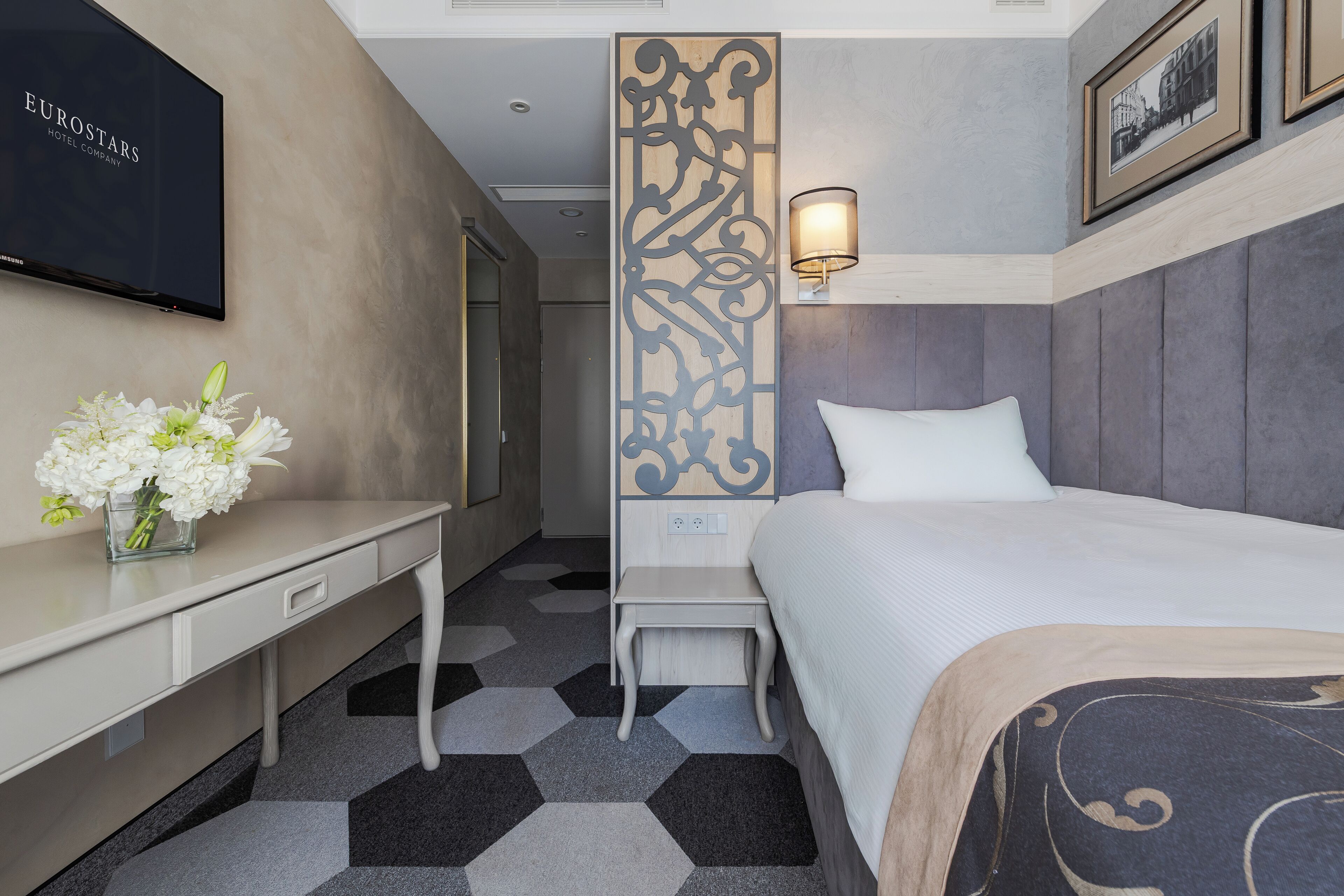 A hotel room with a bed, a desk, a TV, a decorative panel, and a nightstand with a lamp.
