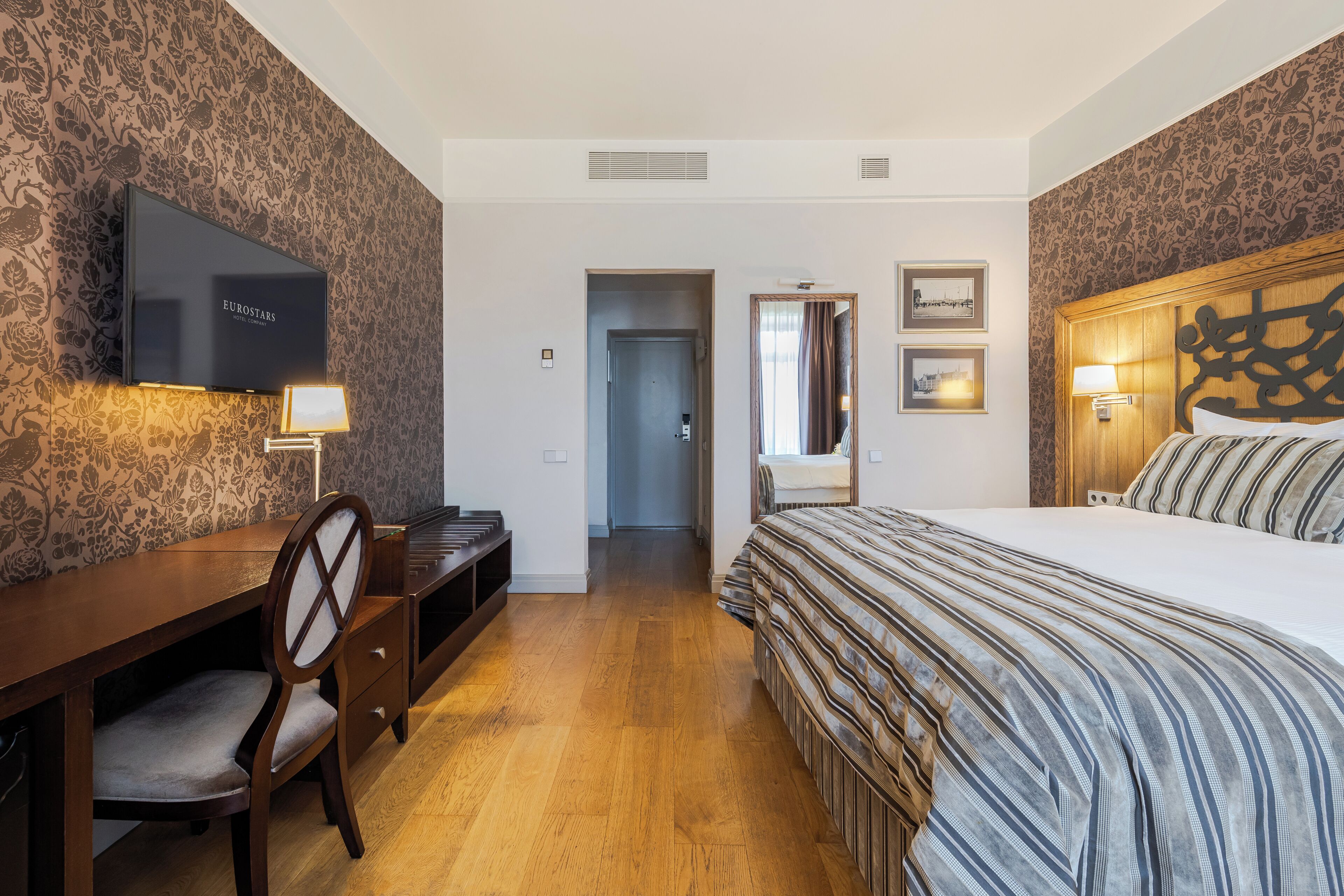 A hotel room with a bed, a desk with a chair, a television, and framed pictures on the wall.
