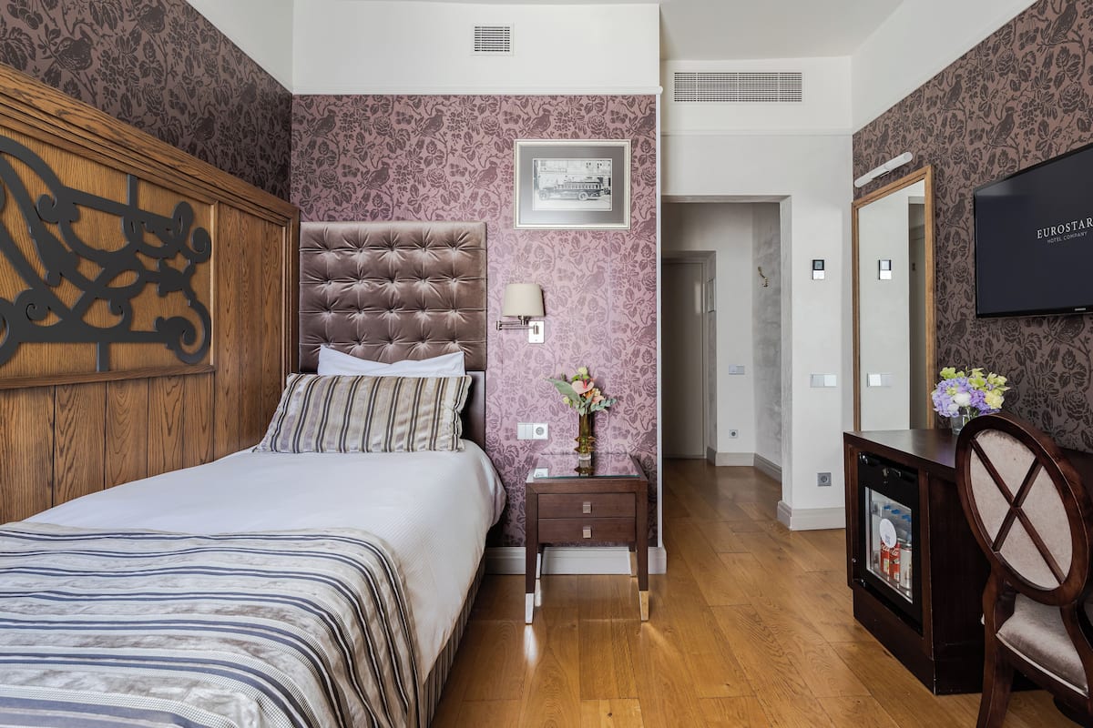 A bedroom with a tufted headboard, a bed with striped bedding, a nightstand with a vase, a mirror, and a television on the wall.