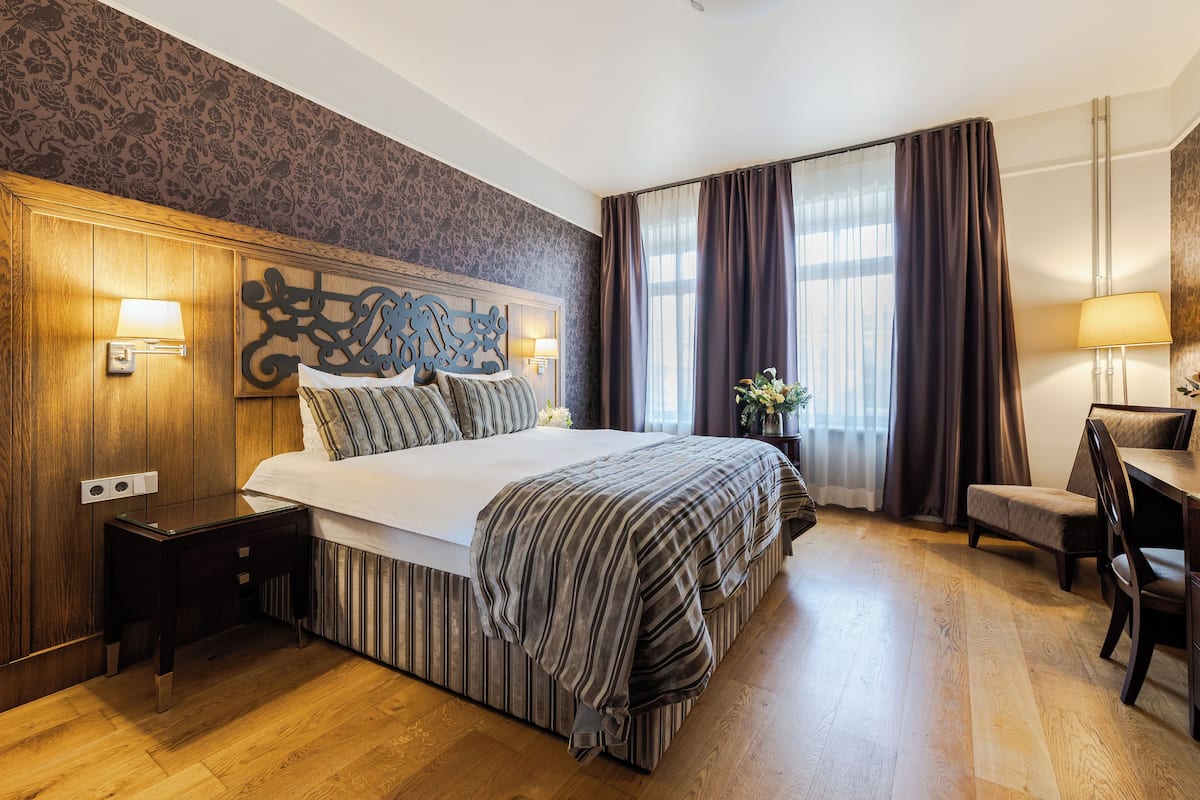 A hotel room with a large bed, wooden headboard, a small desk with a chair, and a window with curtains.