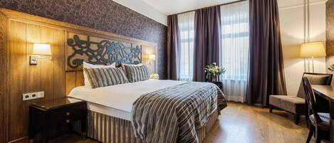 Habitación de hotel con una cama grande, cabecero de madera, un escritorio pequeño con una silla y una ventana con cortinas.