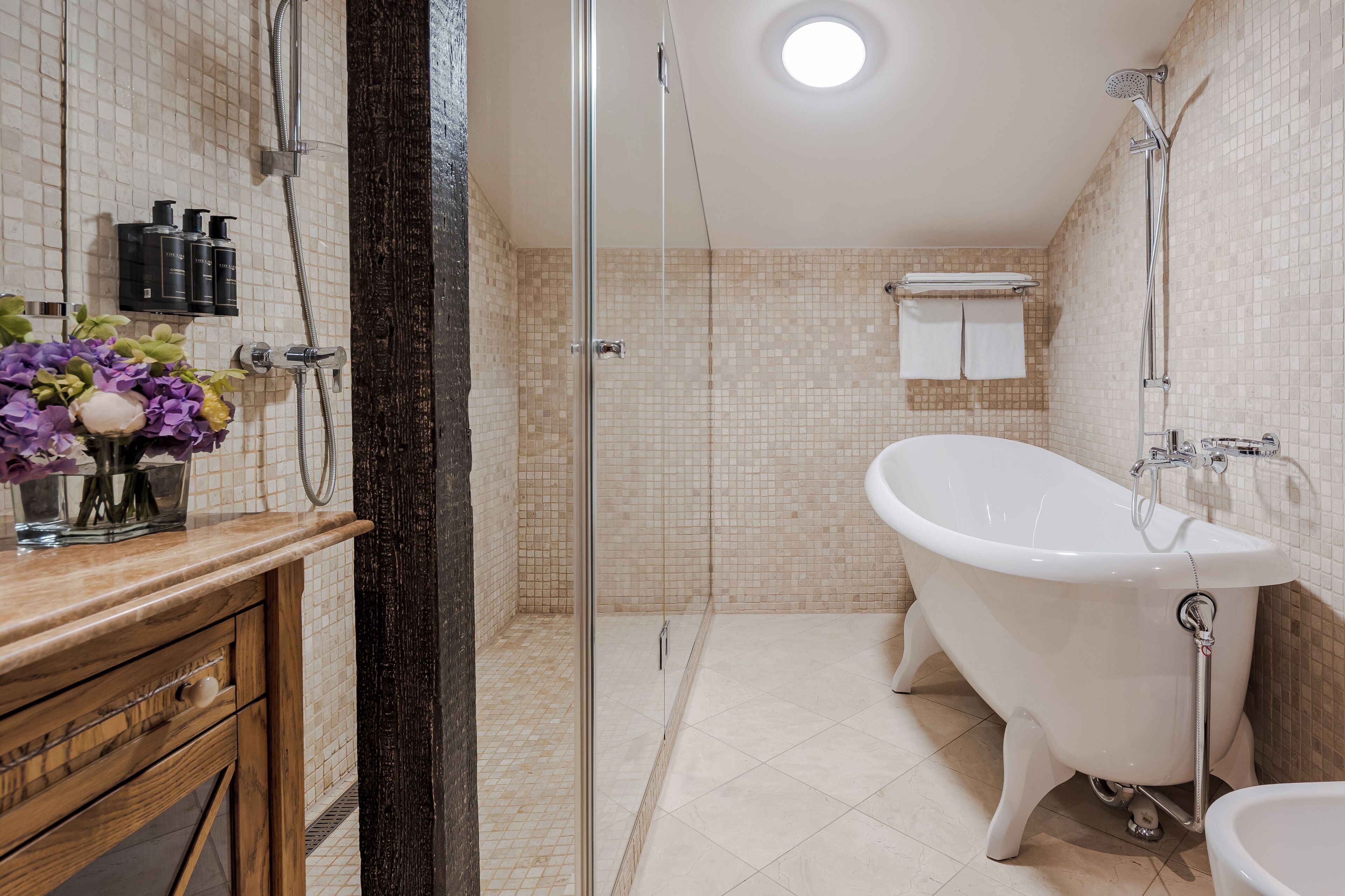Ein Badezimmer mit freistehender Badewanne, einer Duschkabine aus Glas und einem hölzernen Waschtisch mit Blumen in einer Vase.