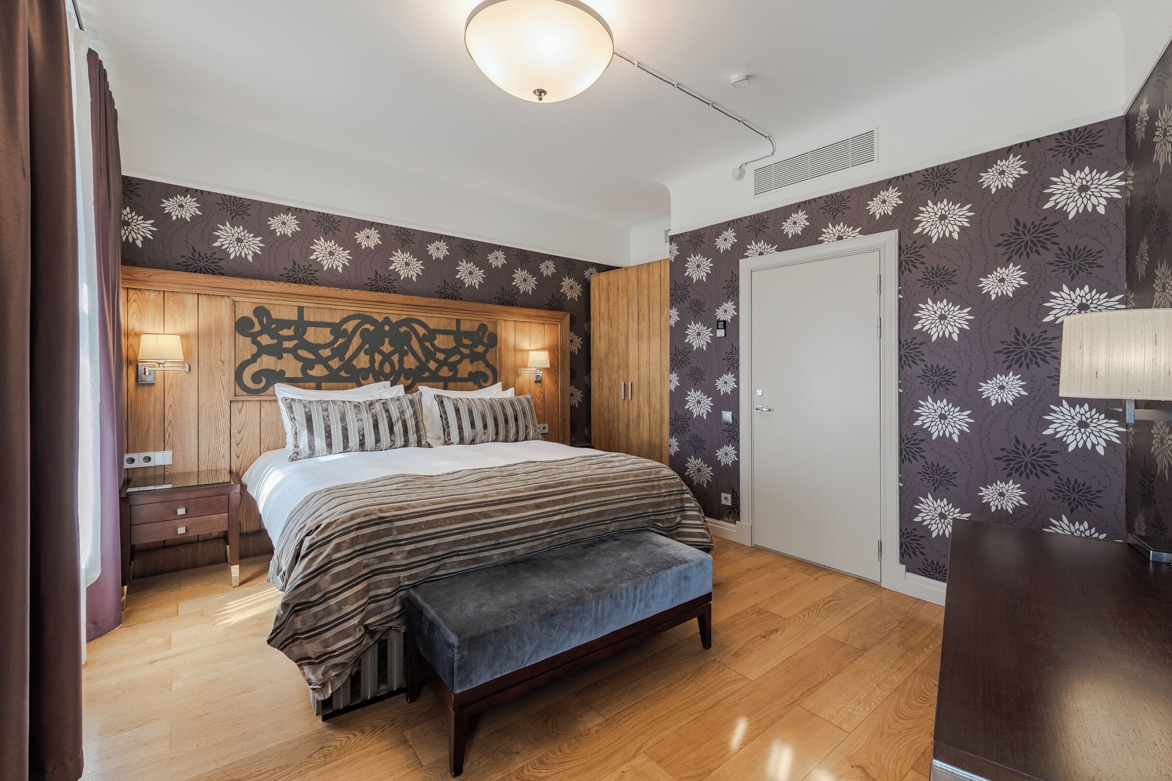 Un dormitorio con cabecera de madera, cama con ropa de cama a rayas, un banco, una mesita de noche y una lámpara de pared.