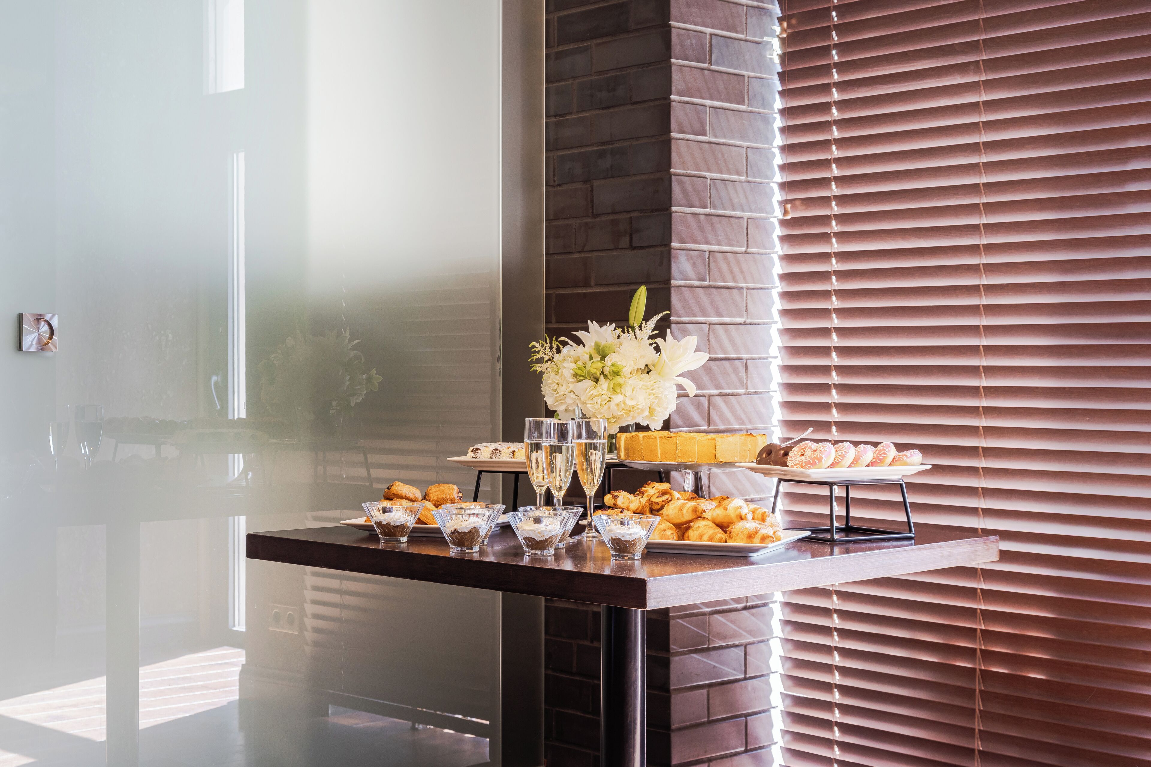 A buffet table with various food items and drinks, set against a brick wall and blinds.