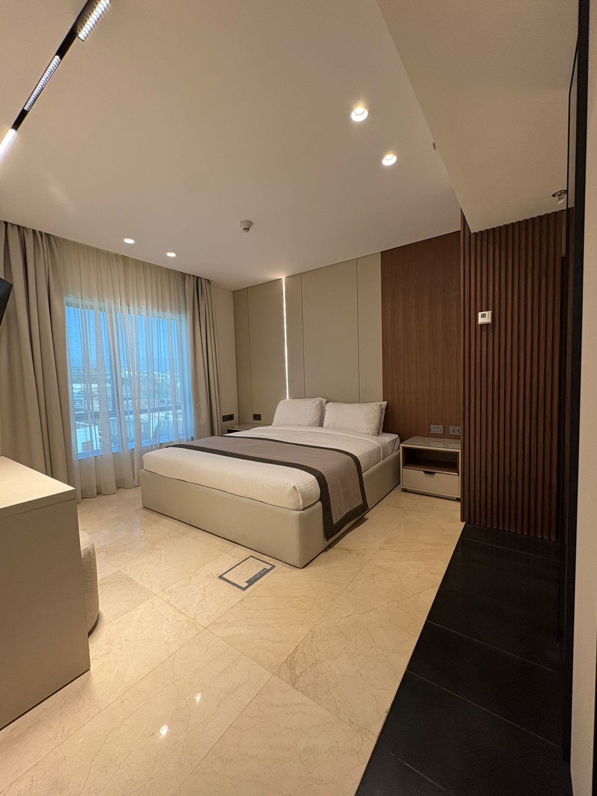 A modern bedroom with a large bed, a bedside table, a nightstand, and a view of the city through the window.