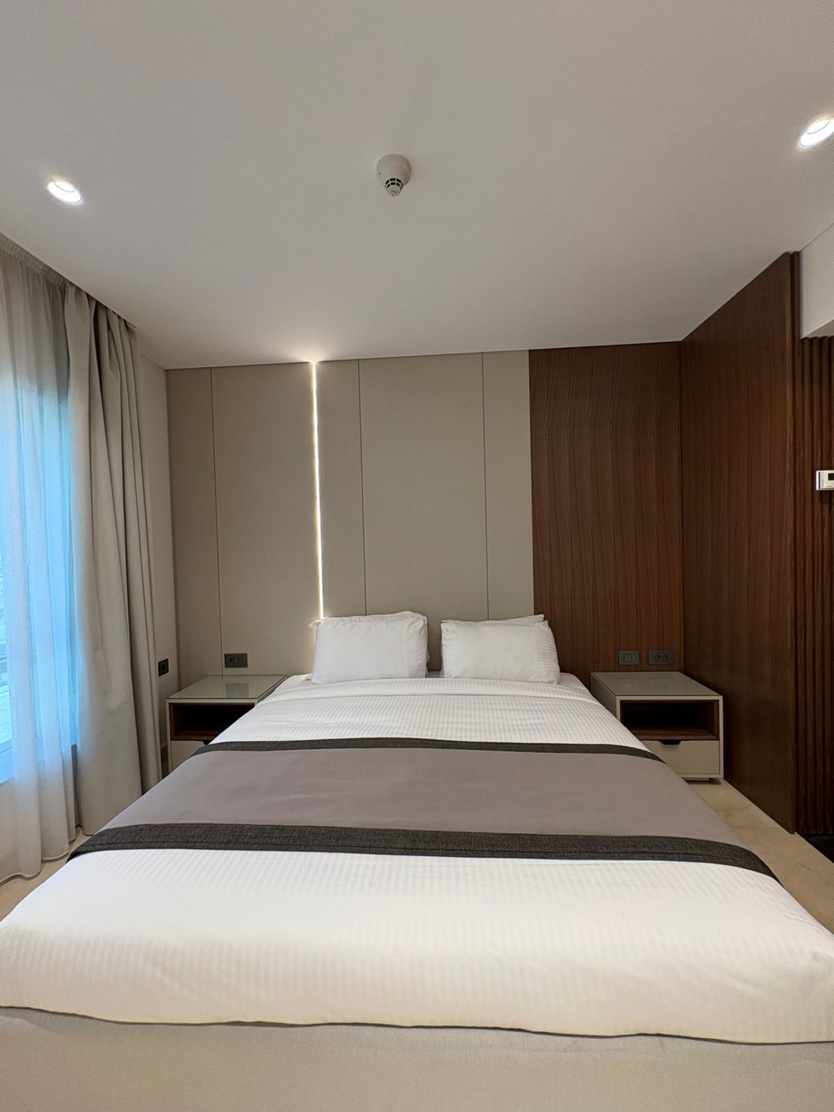 Una habitación de hotel moderna con una cama grande, mesitas de noche y un detector de humo en el techo.