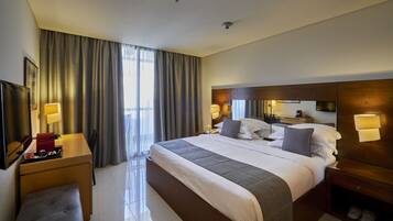 A hotel room with a large bed, bedside tables, a TV, a desk, and a window with curtains.