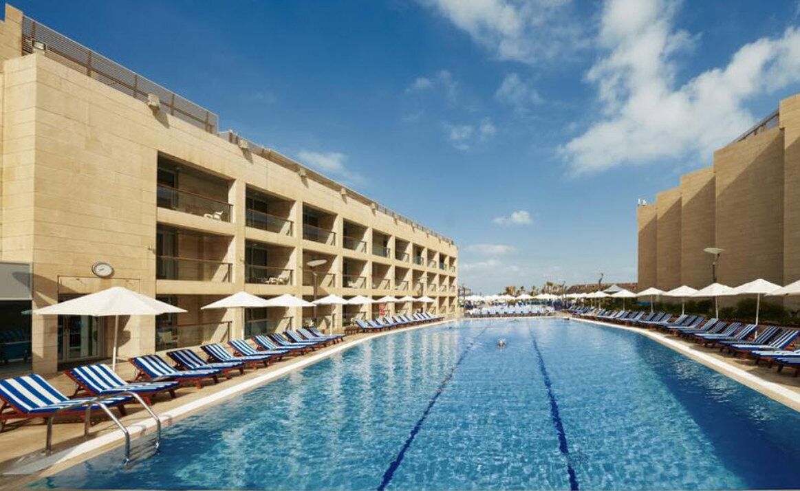 Una piscina con sillas de descanso y sombrillas, rodeada por un edificio de varios pisos y balcones.