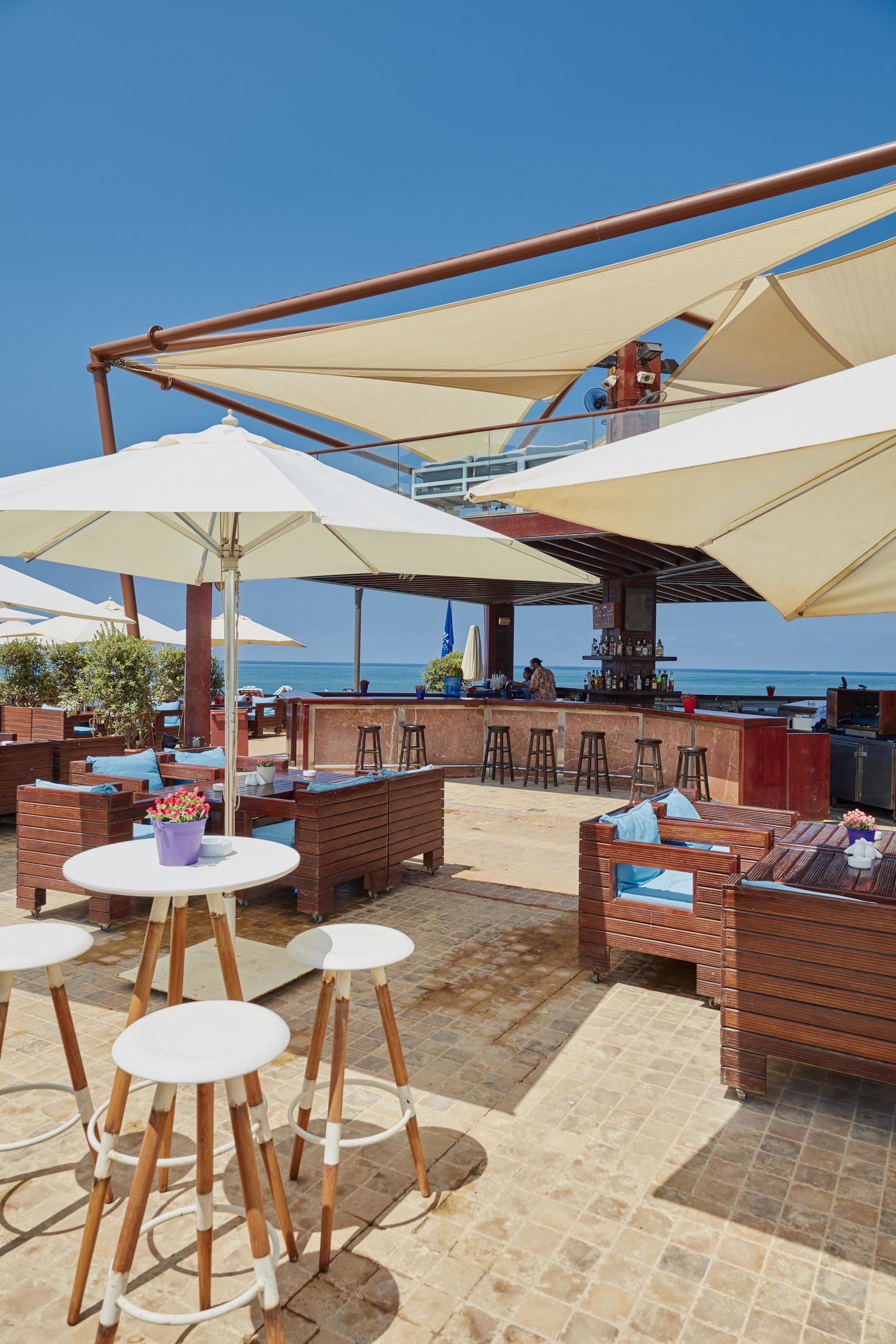 An outdoor seating area with wooden furniture and white tables, a bar with stools, and large umbrellas providing shade.