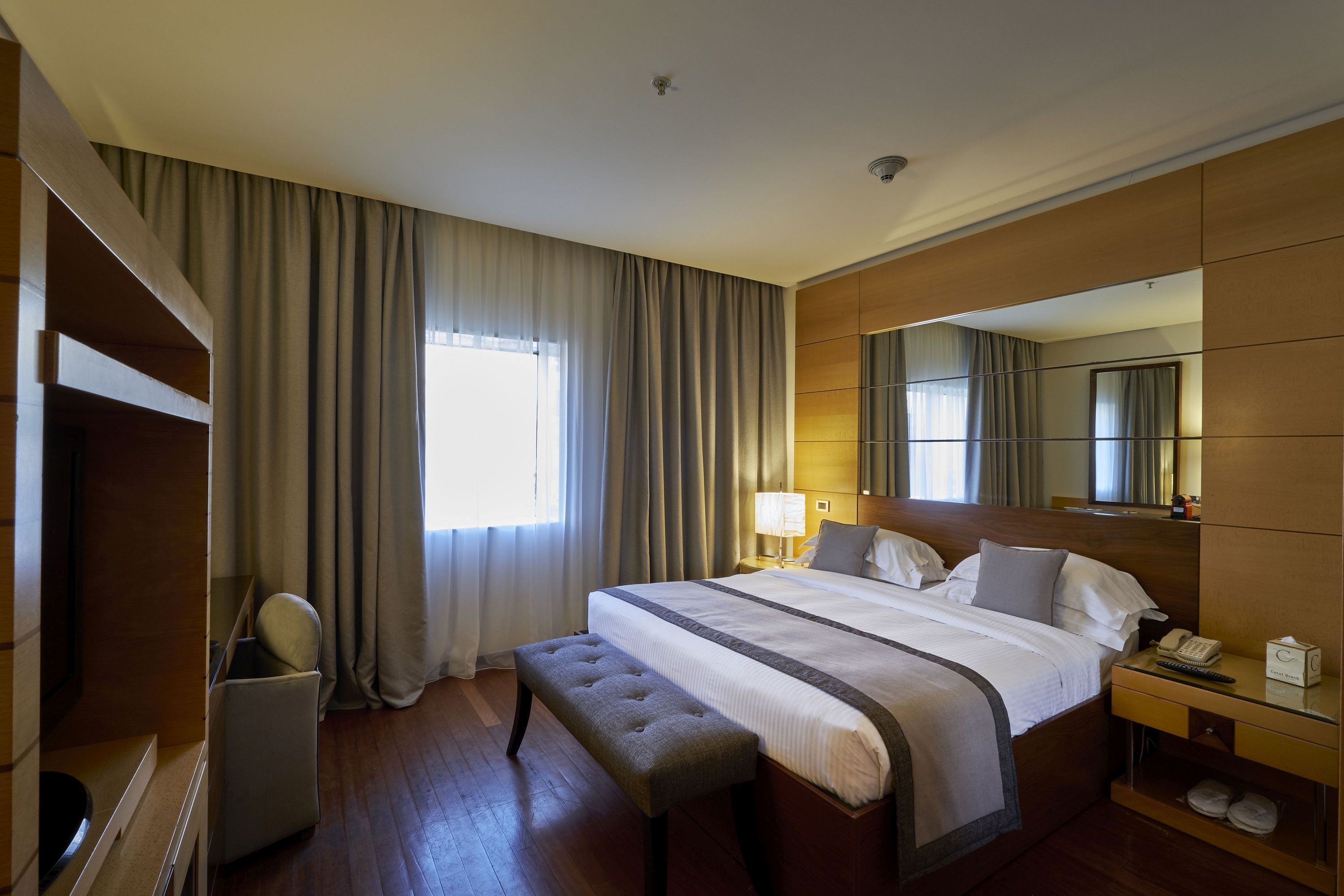 A hotel room with a large bed, a bedside table with a lamp, a bench, a wardrobe, and a mirror.