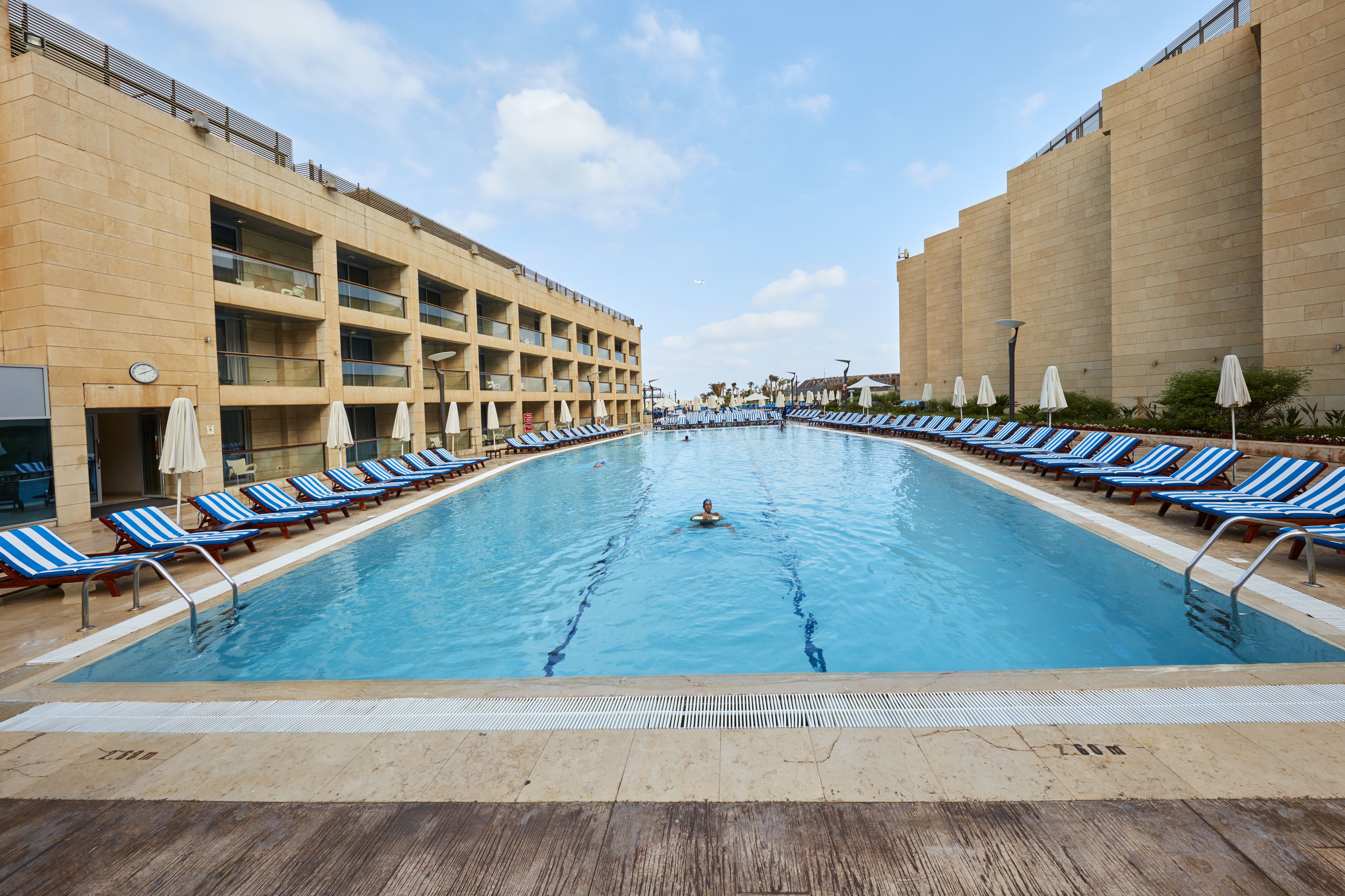 Una piscina con sillones y sombrillas a rayas azules y blancas, rodeada por un edificio grande con varios balcones.